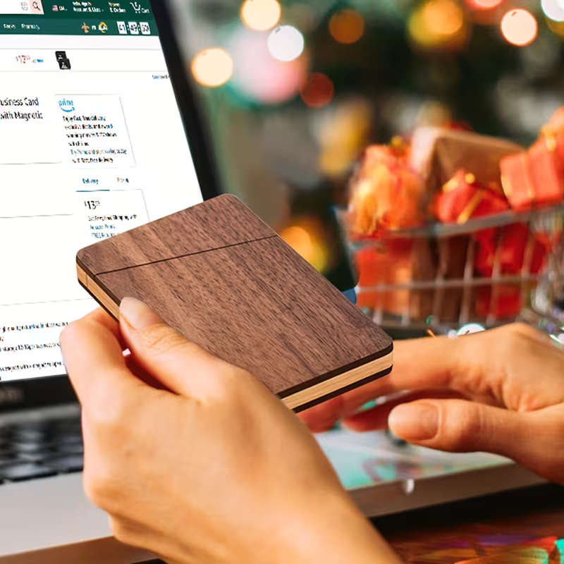 Personalized wood-grain card holder held in hands, with holiday gifts and laptop behind it.